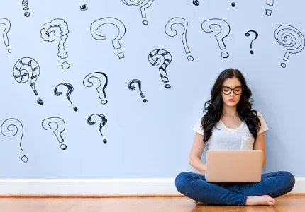Woman sitting on the floor with a laptop, surrounded by question marks on a light blue wall.